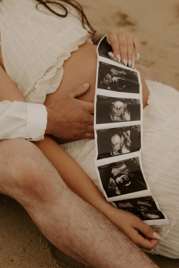 Couple holding ultrasound photos over the mother’s baby bump during a beach maternity photoshoot.