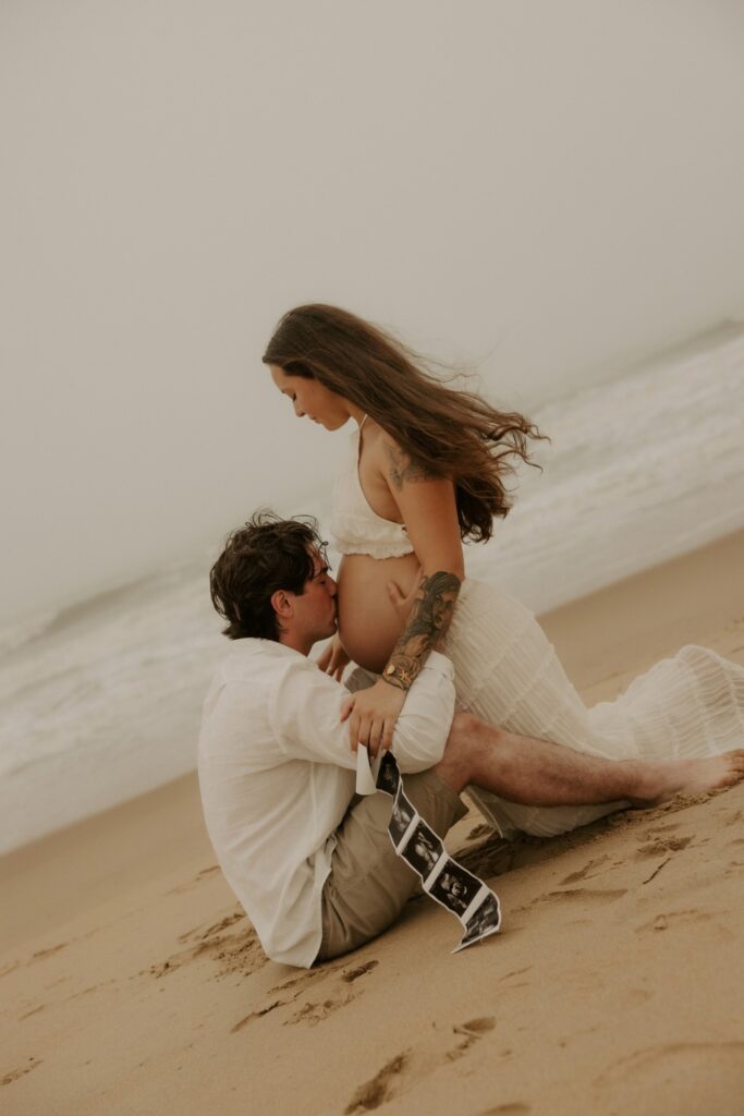 Partner kissing the expectant mother’s baby bump while sitting together on the beach.