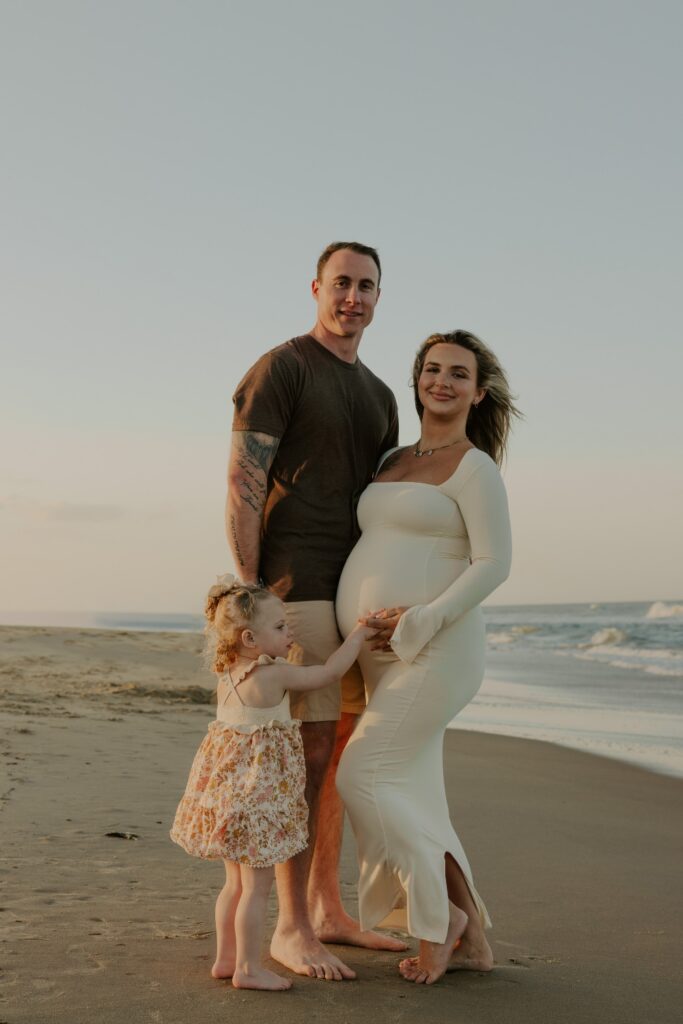 Expectant parents posing barefoot with their daughter beside the ocean at golden hour.