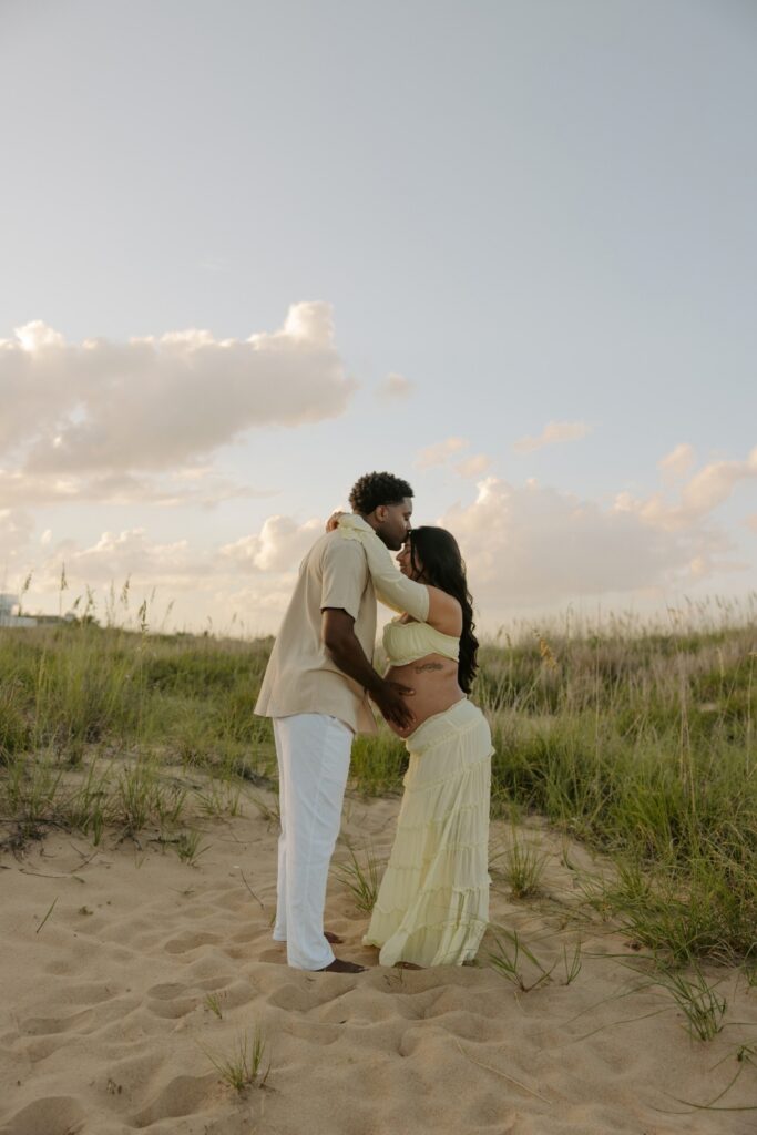 Couple embracing beside the beach dunes during their golden-hour maternity photoshoot.