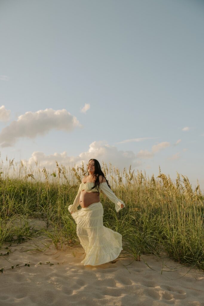 Expectant mother walking through beach dunes in a flowing neutral maternity outfit.