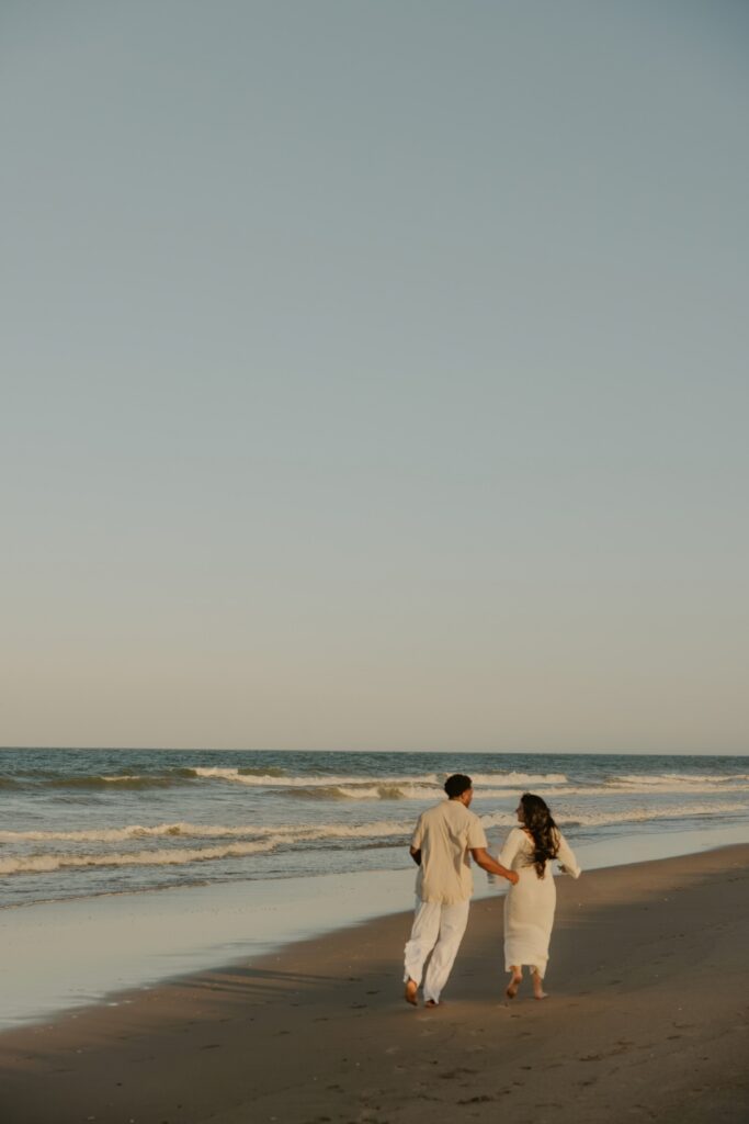 Expectant couple running barefoot along the shoreline during a beach maternity photoshoot.